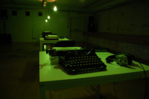 A row of green typewriters stretches off into the distance underneath a low of bare lightbulbs