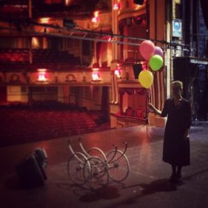 A woman stands on an empty stage, holding a bunch of balloons. We see a grand, empty auditorium behind her.